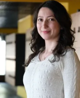 A woman with long, dark, wavy hair stands indoors, facing the camera with a slight smile. The background is softly blurred, with yellow, black, and neutral architectural elements.  